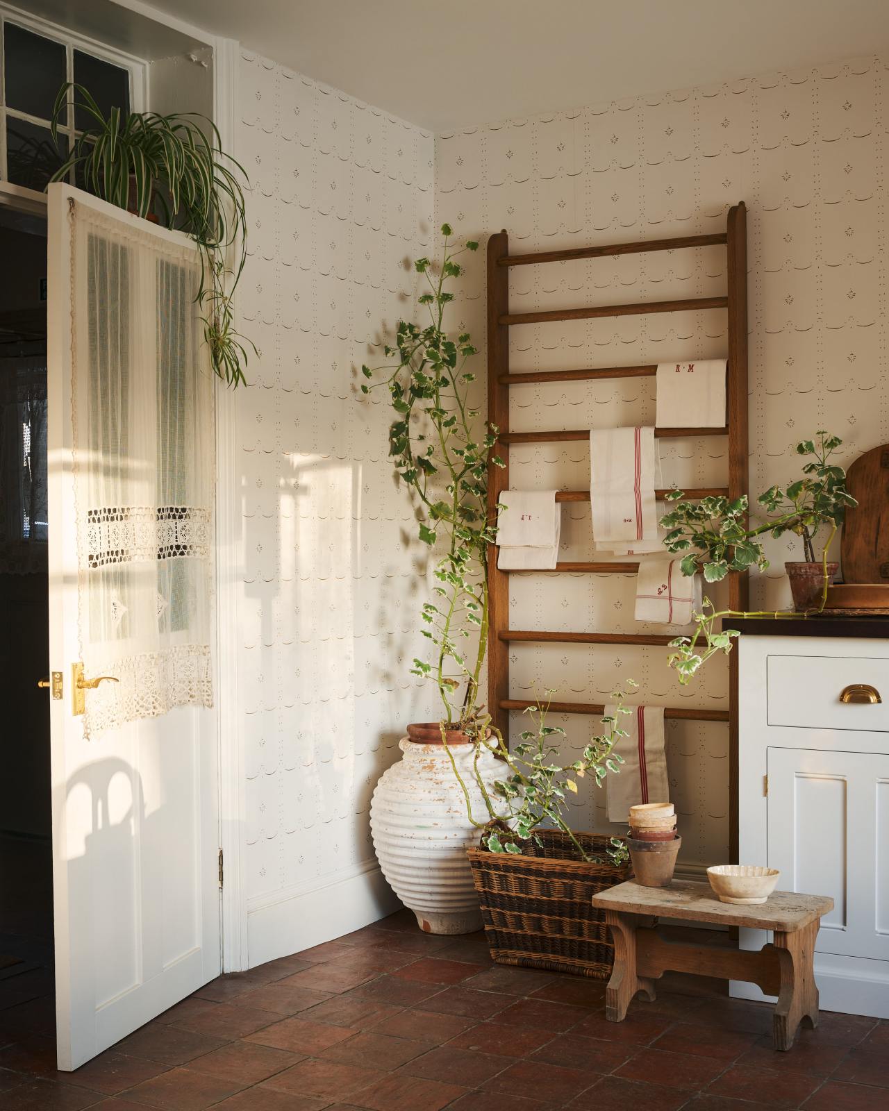 A corner of a Mediterranean-inspired room with a terracotta floor, light tones and an off-white wallpaper with a delicate sepia-coloured pattern.