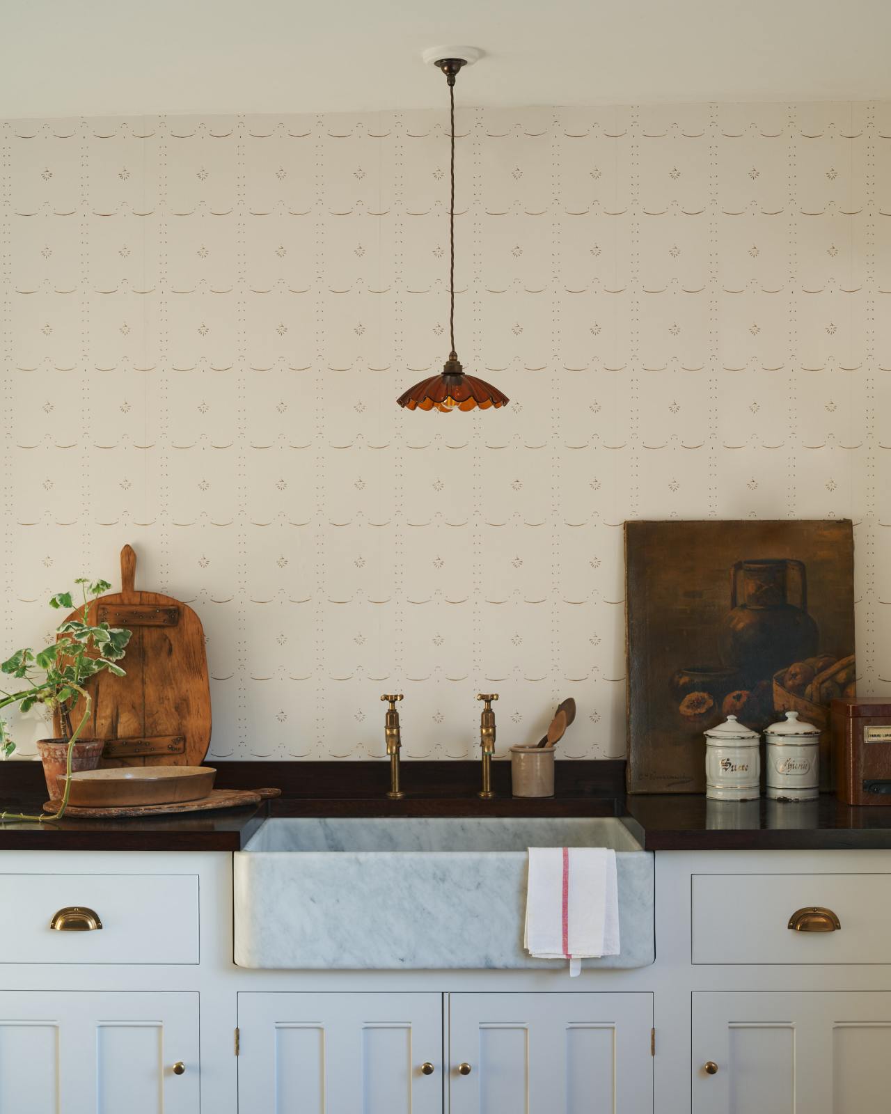 An off-white wallpaper with sepia details behind a white kitchen cabinet with a marble sink and dark wooden worktops.