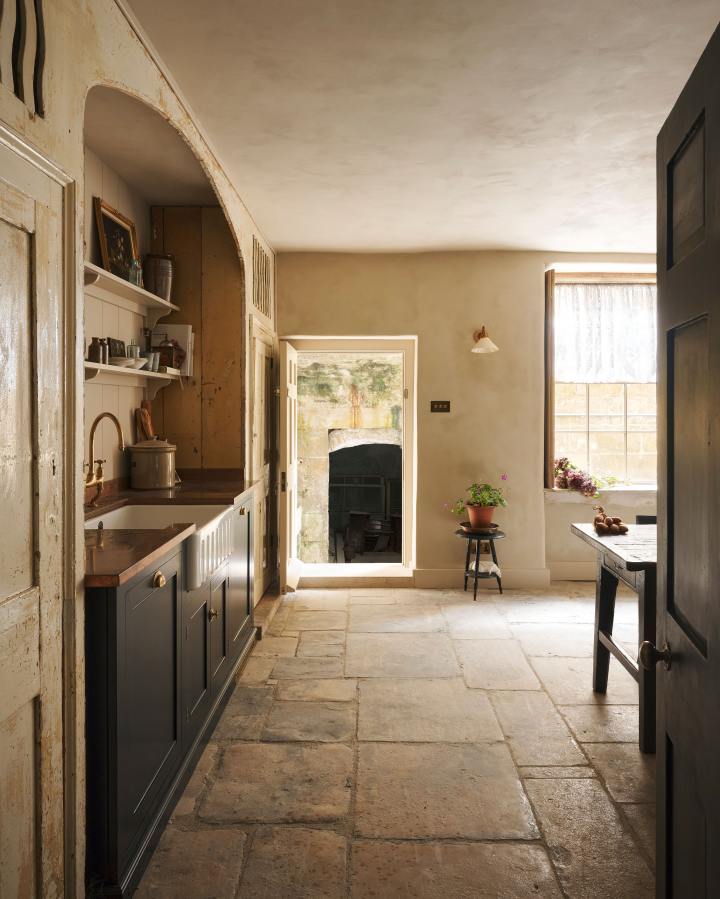 Looking through the entranceway to a rustic basement kitchen with sandy-coloured walls and ceiling, chipped paintwork and a worn stone floor. On the left is a short run of elegant black cupboards with a soft sheen and a mottled copper worktop. Ahead, light pours into the room through a large window with original wavy Georgian panes and an open doorway leading outside to a stone courtyard.