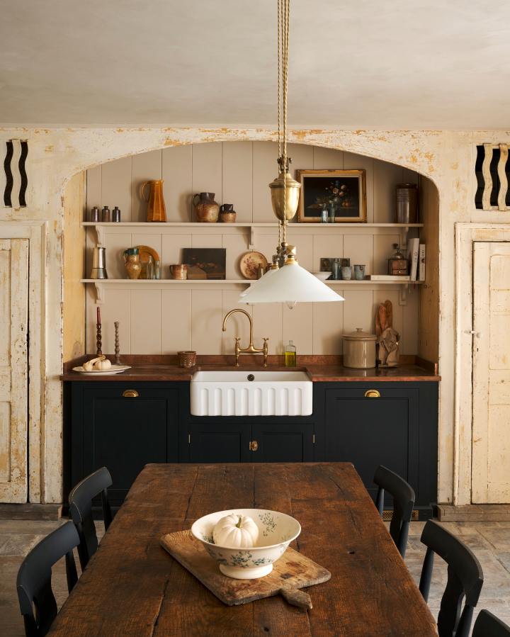 Looking through the entranceway to a rustic basement kitchen with sandy-coloured walls and ceiling, chipped paintwork and a worn stone floor. On the left is a short run of elegant black cupboards with a soft sheen and a mottled copper worktop. Ahead, light pours into the room through a large window with original wavy Georgian panes and an open doorway leading outside to a stone courtyard.