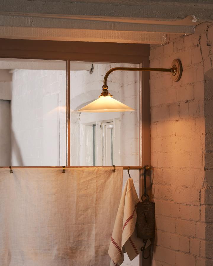 A translucent milk glass shade is suspended from a peach-coloured brick wall by a straight brass arm which curves downwards at a right angle at the end. Warm light glows through the glass shade, illuminating a moody corner space.