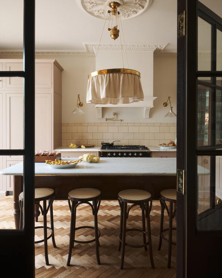 Looking through a glazed partition into a stylish Shaker kitchen with pink painted cupboards, cream wall tiles, a fabric chandelier and a Dairy Table with a row of rattan café stools.