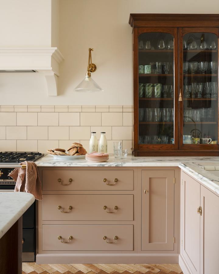 Looking through a glazed partition into a stylish Shaker kitchen with pink painted cupboards, cream wall tiles, a fabric chandelier and a Dairy Table with a row of rattan café stools.