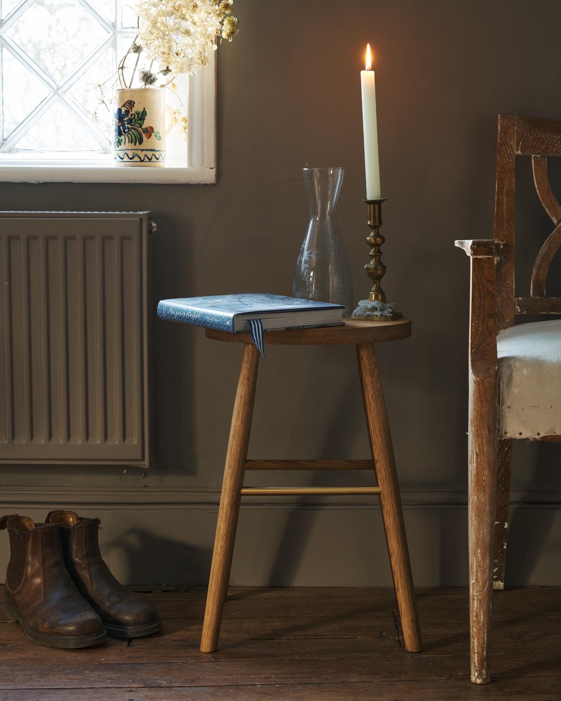 A straight-on view of a simple oak milking stool with an oval-shaped seat and a brass footrail in a dark room. A hardback book and a lit pillar candle are placed ontop of the stool.