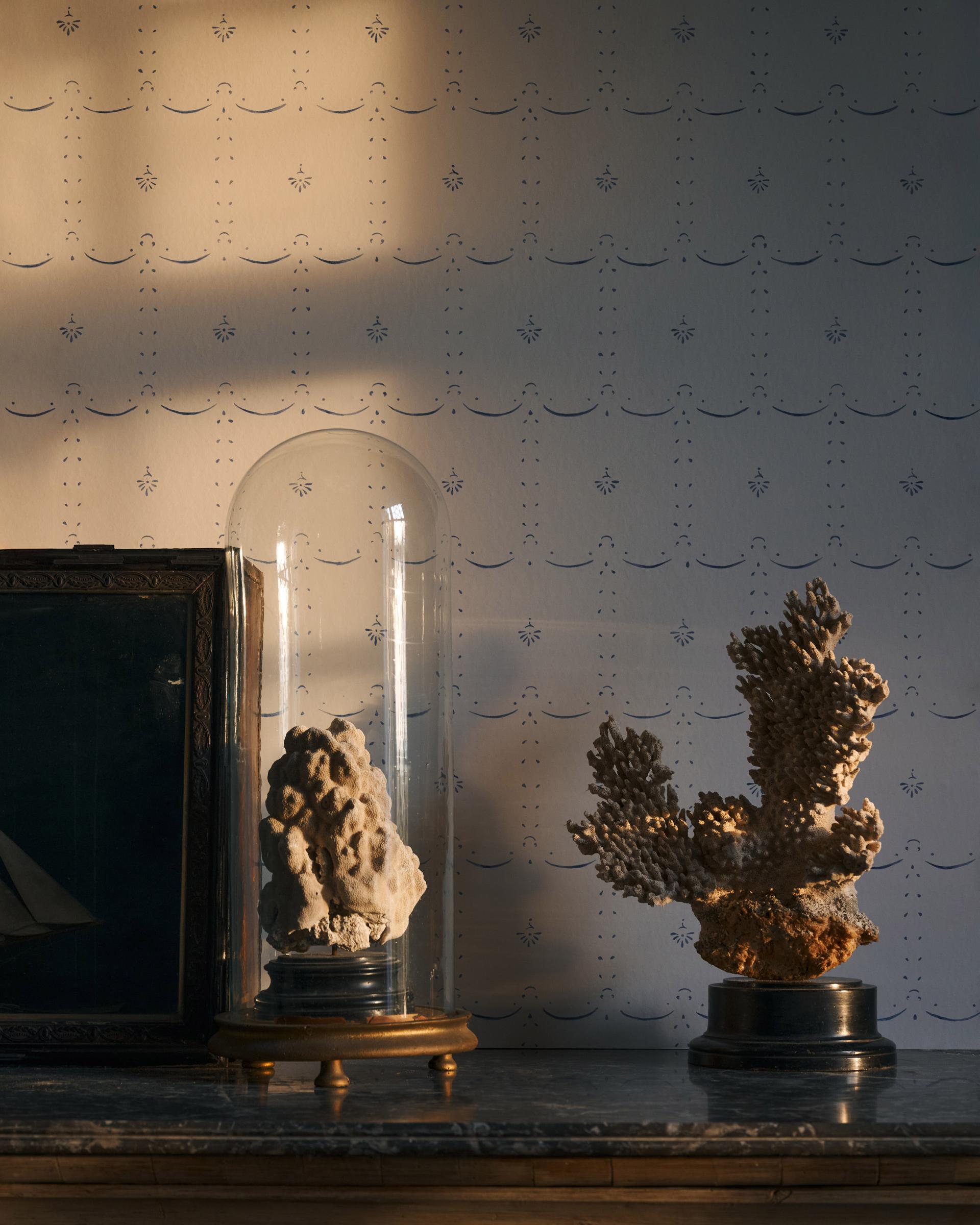 Warm light from a sunset is falling across a delicate patterned wallpaper with the shadow of a window frame visible behind antique ornaments on a table.