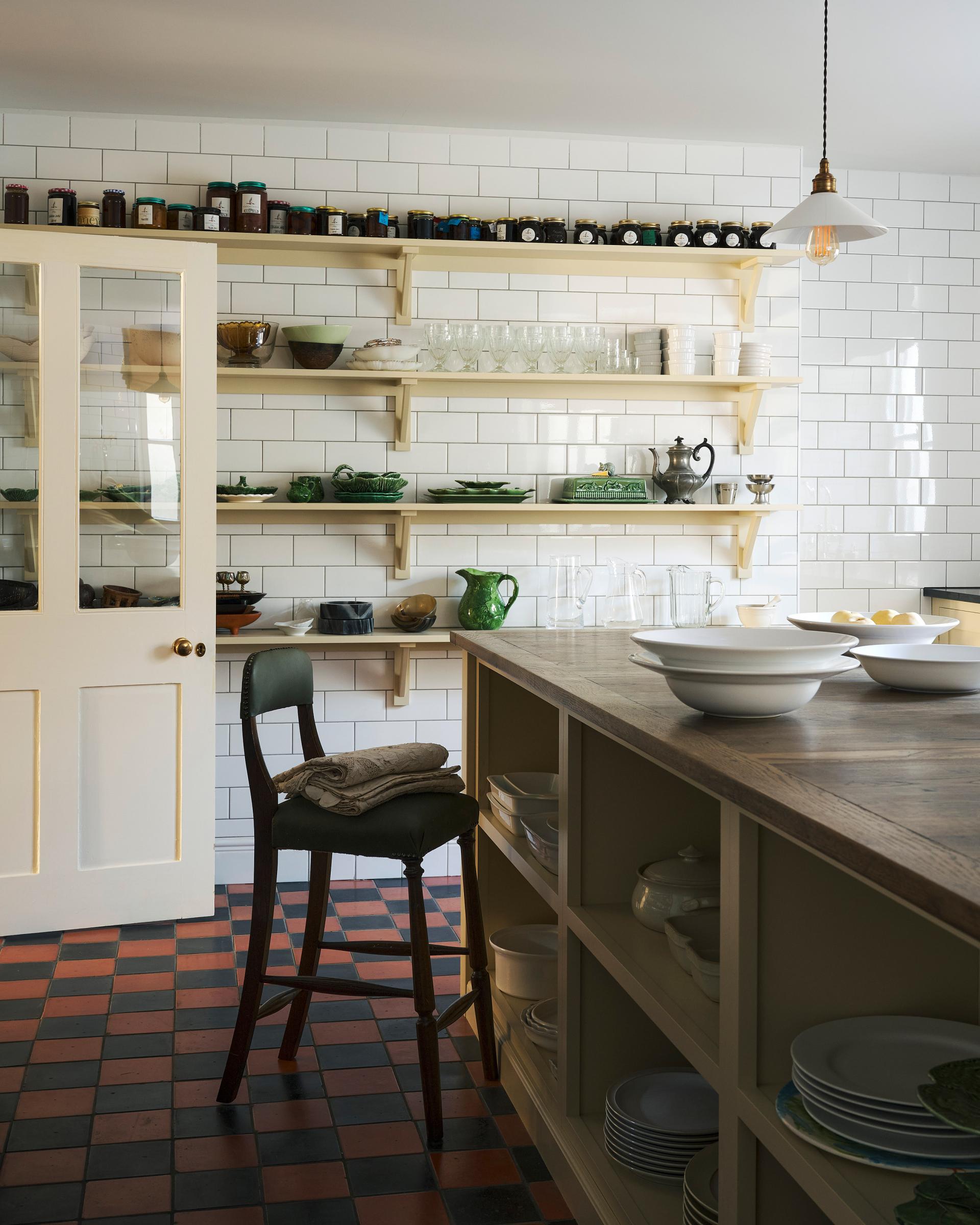 An atmospheric shot of an antique green leather upholstered bar stool, propped up at a large Shaker island in the centre of a utilitarian-looking scullery. Behind the island, four rows of Shaker shelves, painted in a pale yellow hue, are fitted to a wall of glossy white subway tiles, a soft contrast to the original red and black chequerboard quarry tile floor.