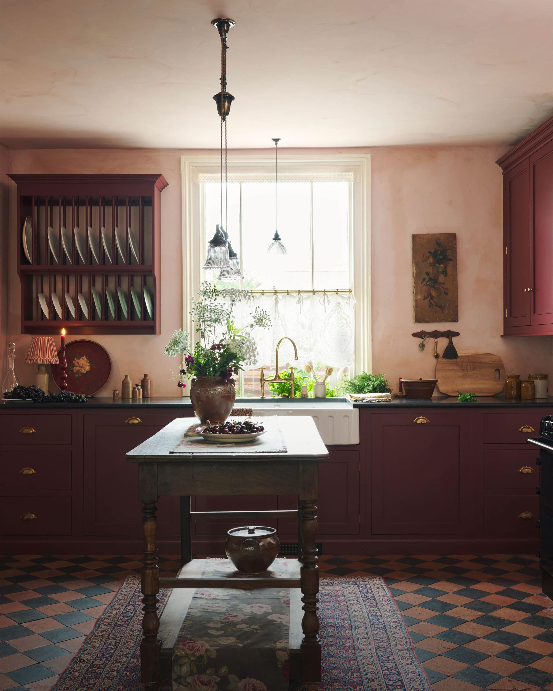 A warm and simply gorgeous refectory-red Shaker kitchen features a sink run with a black granite worktop, a wall cupboard, a small Heirloom plate rack, a pantry cupboard, and a gas stove in a chimney breast with Sepia Lace market splashback tiles. Beautifully decorated with vintage furniture, artisan ceramics, original botanical illustrations, reeded glass pendant lights, and a wild floral arrangement, it creates a humble and endearing space.