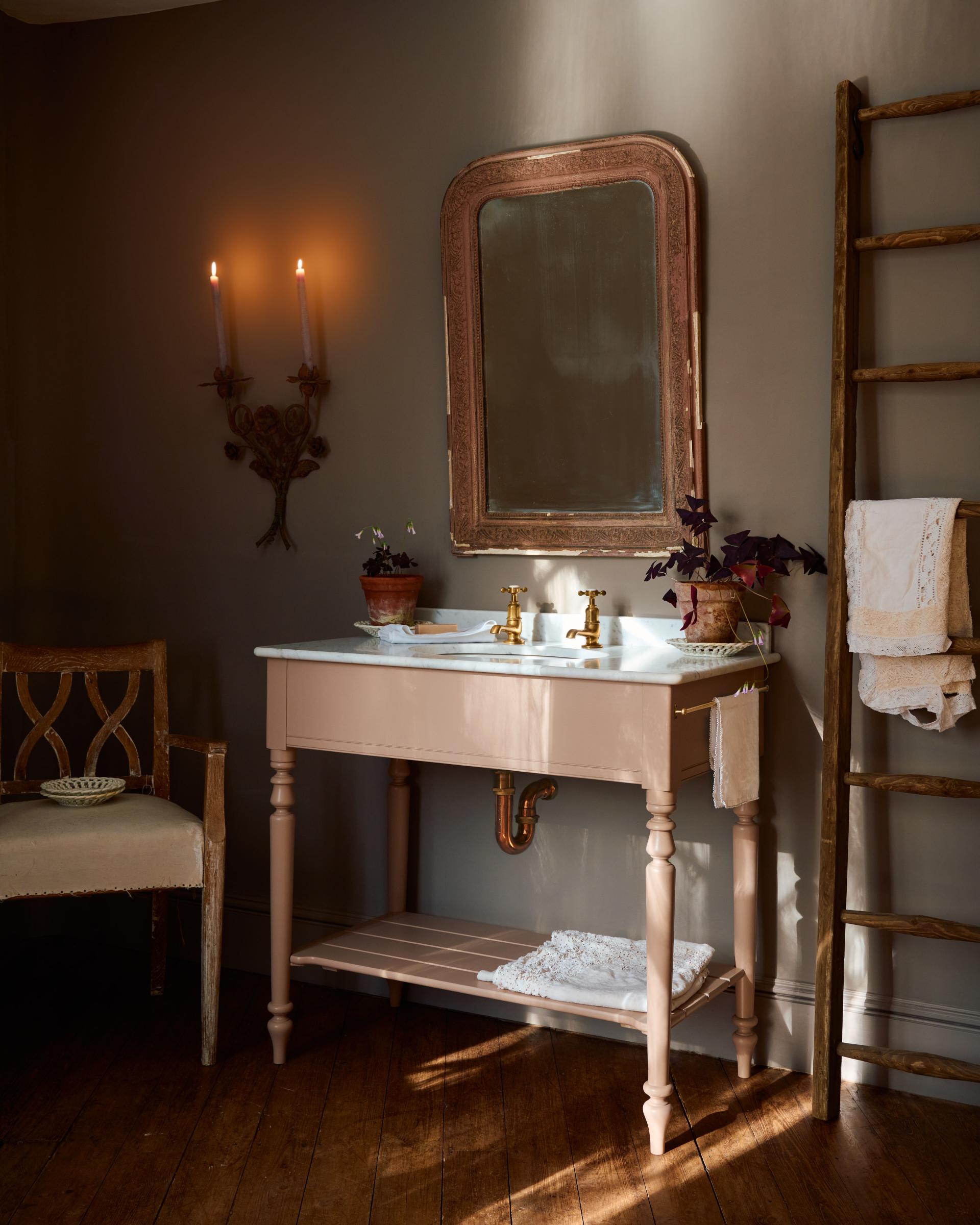 An oblique view towards a dusty-pink Victorian-inspired washstand with brass pillar taps in a greyish-brown painted room. The shot is atmospheric and moody, with two lit candles casting a subtle glow and light from an adjacent window illuminating the brass accents.
